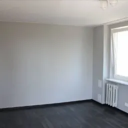 Pomalowane na szaro puste pomieszczenie z ciemną podłogą, białym grzejnikiem pod oknem i widocznymi puszkami elektrycznymi w ścianie.