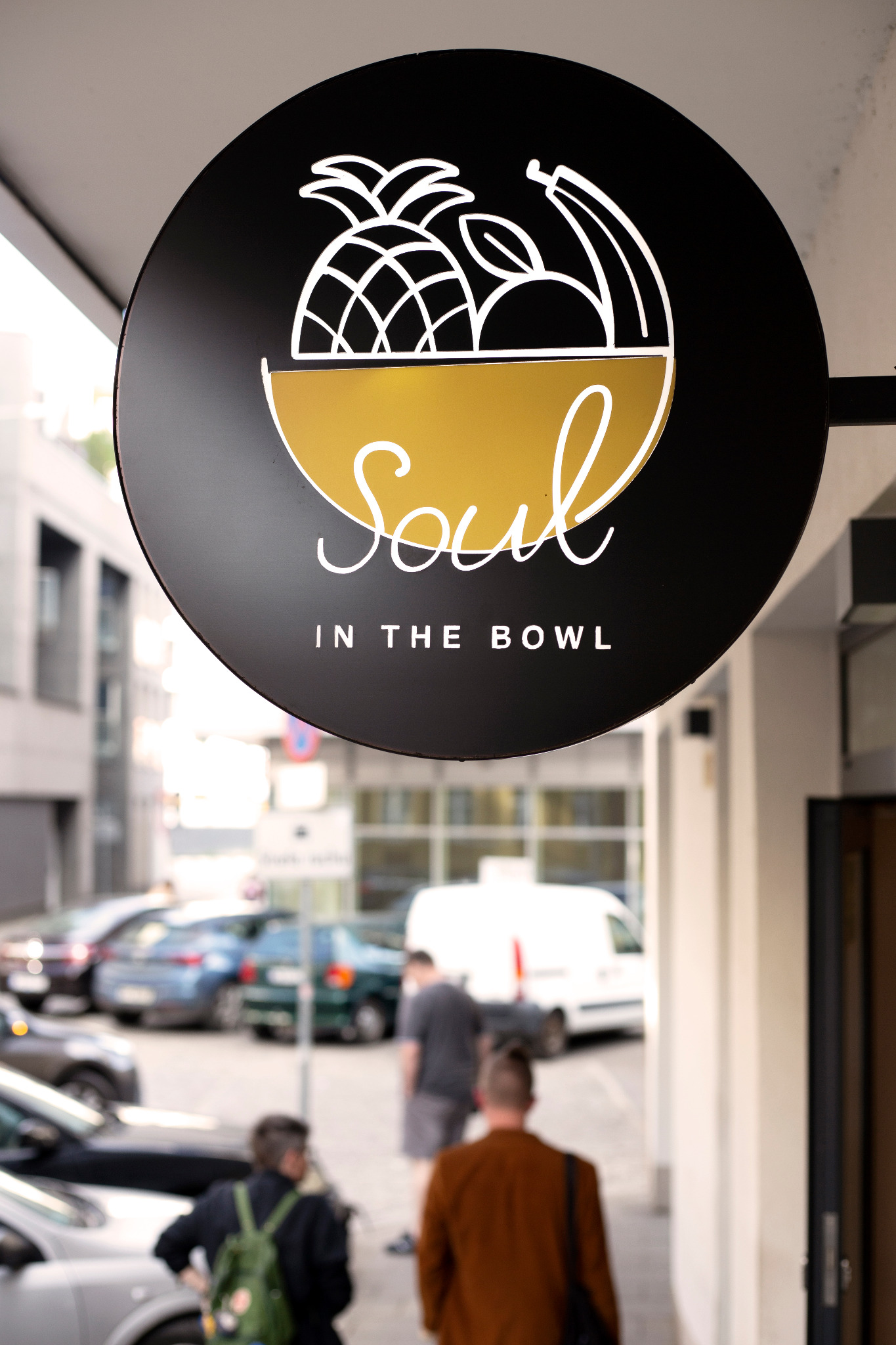 Okrągły szyld kawiarni SOUL IN THE BOWL z grafiką owoców w misce, zawieszony na zewnątrz budynku, widok z perspektywy przechodnia na ulicy.