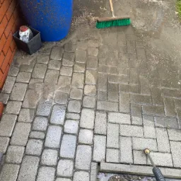 Częściowo umyta kostka brukowa z widoczną różnicą w czystości, niebieski pojemnik, szczotka i lanca myjki ciśnieniowej