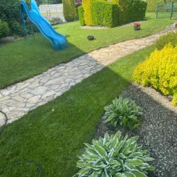 Uporządkowany ogród z równo przystrzyżonym trawnikiem, ścieżką z nieregularnych kamieni, formowanymi krzewami i niebieską zjeżdżalnią dla dzieci.