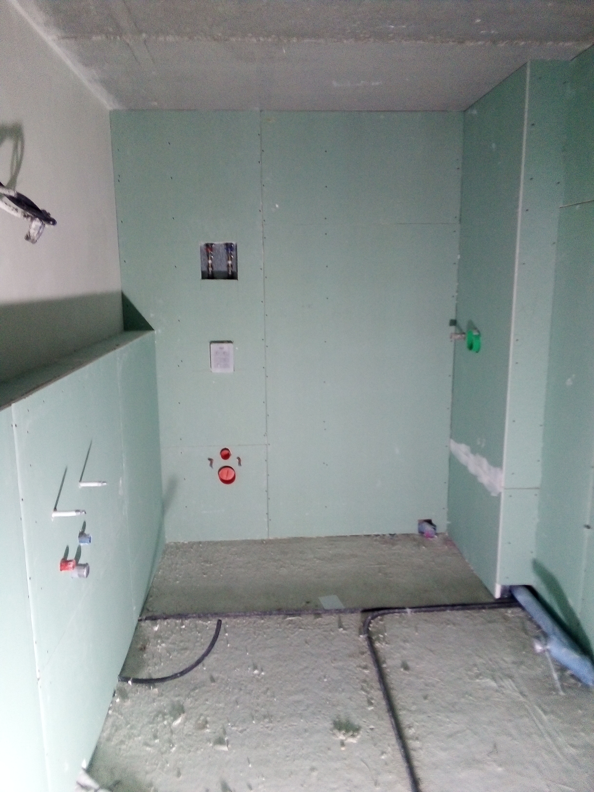 Łazienka w trakcie remontu, ściany obłożone płytami gipsowo-kartonowymi, widoczne instalacje wodne i elektryczne, surowy beton na podłodze.