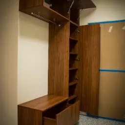 Drewniana szafa wnękowa z otwartymi drzwiczkami i szufladą, widoczne mechanizmy podnoszenia frontów, surowy karton na ścianie w tle.