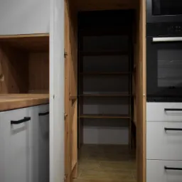 Otwarta spiżarnia z drewnianymi półkami w zabudowie kuchennej z szarymi frontami i drewnianym blatem, widoczny piekarnik i płyta w zabudowie, podłoga wyłożona płytkami imitującymi drewno.