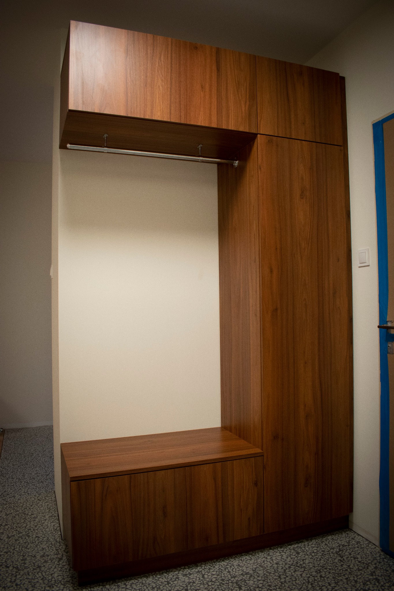 Zabudowa przedpokoju z drewna orzechowego z siedziskiem, wieszakiem na ubrania i szafką, widok na szarą podłogę i niebieskie zabezpieczenie futryny drzwi.