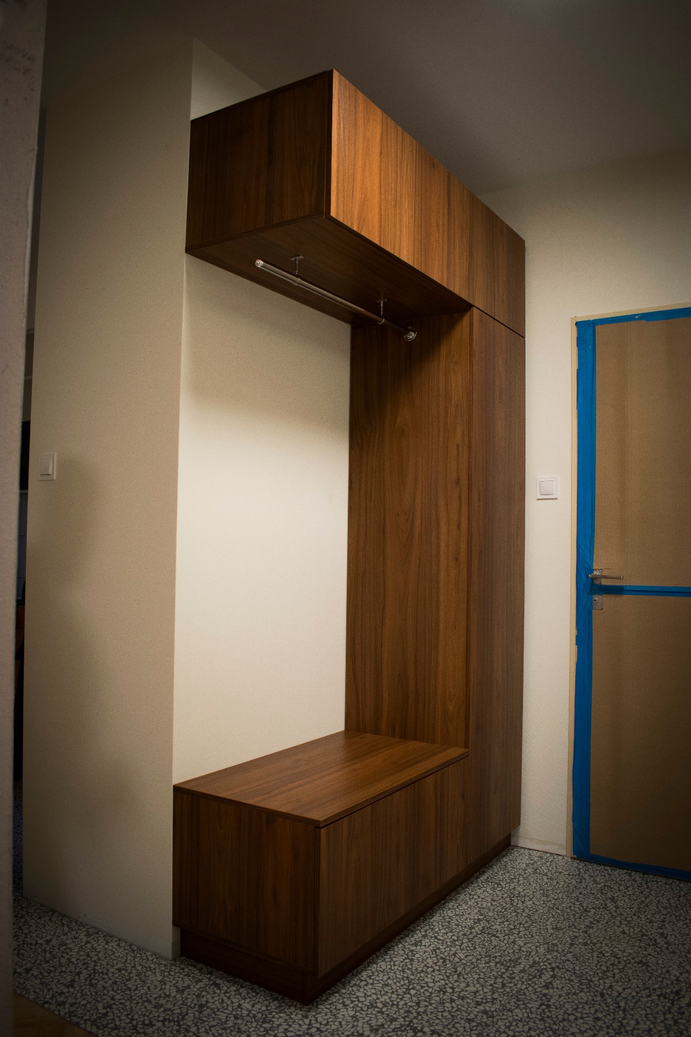 Drewniany przedpokój z szafką, siedziskiem i wieszakiem, wykonany na zamówienie, na tle ściany i drzwi zabezpieczonych taśmą malarską.