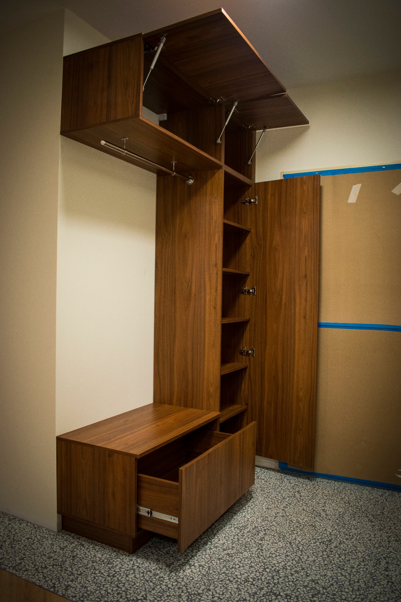 Drewniana szafa wnękowa z otwartymi drzwiczkami i szufladą, widoczne mechanizmy podnoszenia frontów, surowy karton na ścianie w tle.