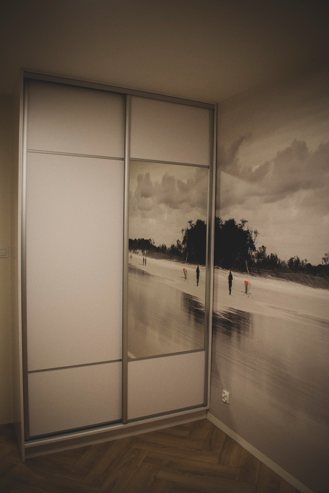 Biała szafa przesuwna z lustrzanymi drzwiami, odbijającymi monochromatyczny widok plaży z postaciami, na tle ściany z fototapetą o podobnym motywie, uzupełniona parkietem w jodełkę.