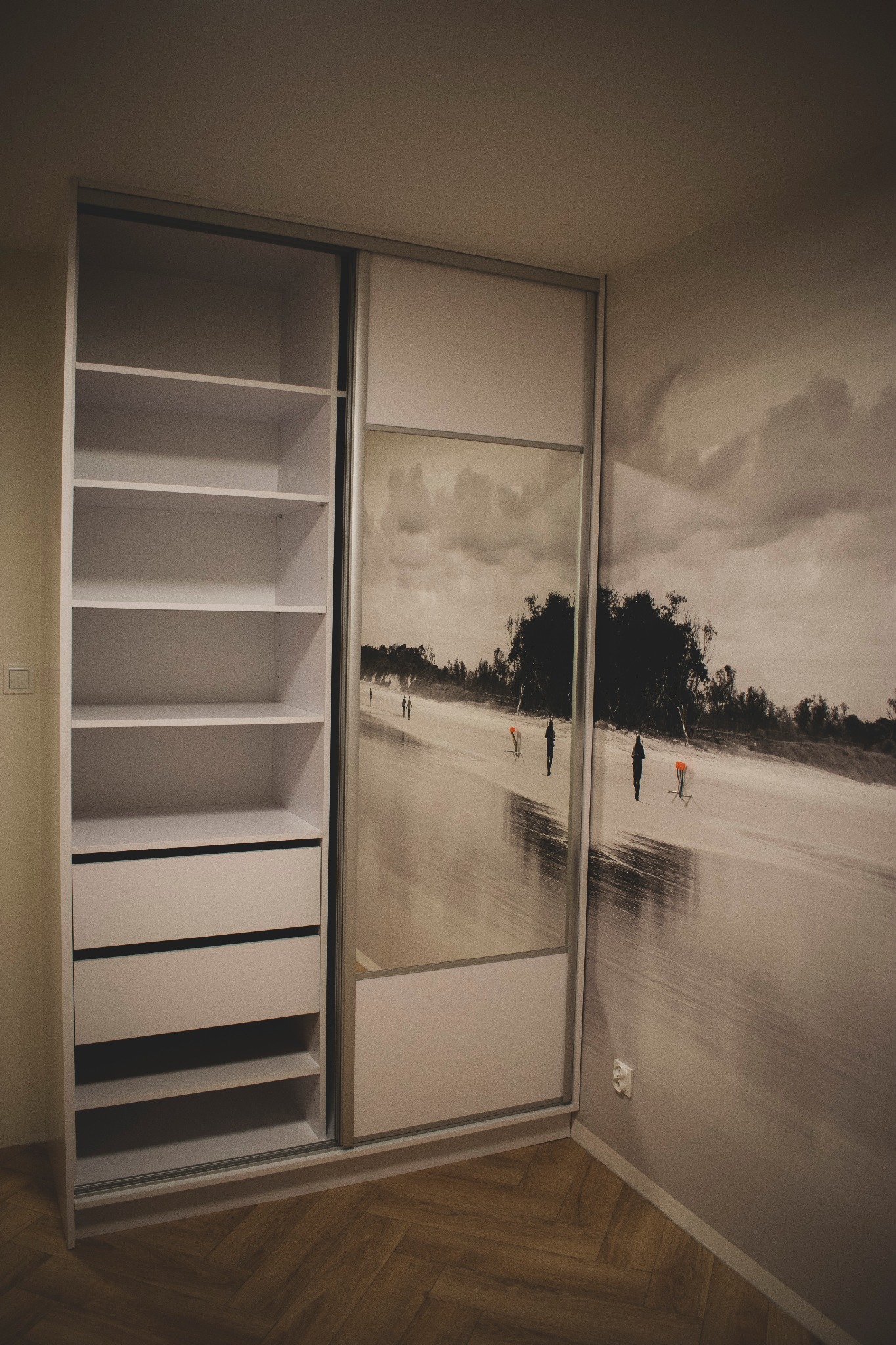 Biała szafa wnękowa z przesuwnymi drzwiami, z lustrem odbijającym plażowy pejzaż, dopasowana do fototapety na ścianie, widok z boku na otwarte półki i szuflady.