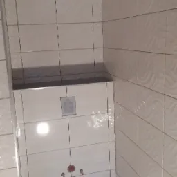 Fragment wykończonej łazienki z beżowymi płytkami o falistej fakturze na ścianach, czarną podłogą i wystającym elementem zabudowy stelaża podtynkowego z widocznymi otworami na rury.