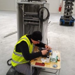 Inneco - Pracownik w kamizelce odblaskowej łączy światłowody przy szafie sterowniczej w hali, drugi monter na podnośniku koszowym w tle.