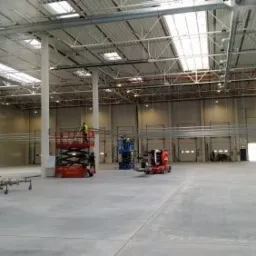 Wnętrze hali magazynowej w trakcie montażu instalacji elektrycznych: widoczne podnośniki koszowe, koryta kablowe pod sufitem i pracownicy w kamizelkach odblaskowych.