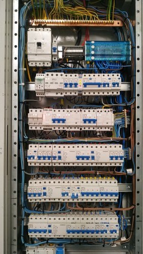 Rozdzielnica elektryczna z licznymi zabezpieczeniami nadprądowymi, okablowaniem w kolorach niebieskim, żółtym i zielonym, oraz szyną wyrównawczą z miedzi. Widoczne elementy automatyki i sterowania.