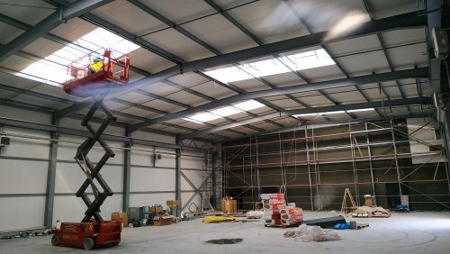 Pracownik w podnośniku nożycowym, wykonujący prace na wysokości wewnątrz hali przemysłowej z widocznymi instalacjami pod sufitem.