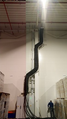 Rozbudowana instalacja kablowa w hali magazynowej, kable prowadzone w korytach pionowo wzdłuż ściany, pracownik przyłącza kolejne przewody do skrzynki rozdzielczej.