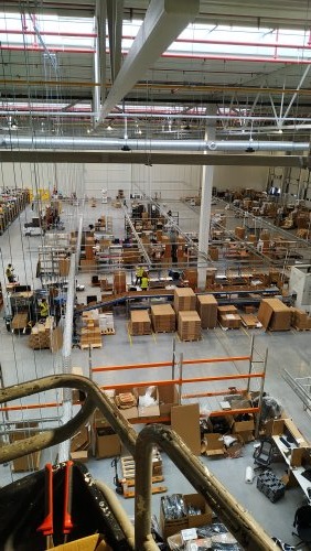 Widok z góry na halę magazynową z widocznymi pracownikami w żółtych kamizelkach, kartonami i regałami; w pierwszym planie fragment metalowej konstrukcji i narzędzia.