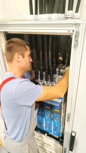 Mężczyzna w kombinezonie z szelkami pracuje w szafie rozdzielczej, układając grube, czarne kable w górnej części i podłączając je do niebieskich modułów zabezpieczeń na dole.