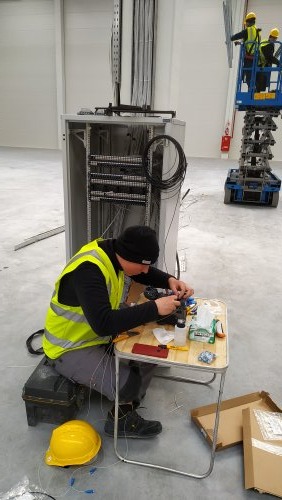 Pracownik w kamizelce odblaskowej łączy światłowody przy szafie sterowniczej w hali, drugi monter na podnośniku koszowym w tle.