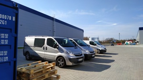 Flota srebrnych samochodów dostawczych zaparkowana przed halą magazynową z niebieskim kontenerem po lewej stronie, palety na pierwszym planie.