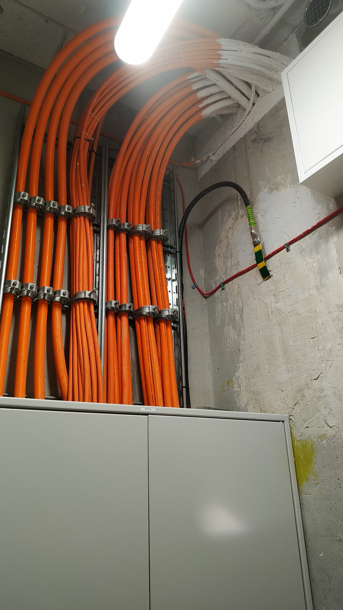 Pomarańczowe przewody elektryczne ułożone równolegle i przymocowane metalowymi uchwytami do ściany, część przewodów pomalowana na biało przy suficie, szara szafa instalacyjna u dołu.