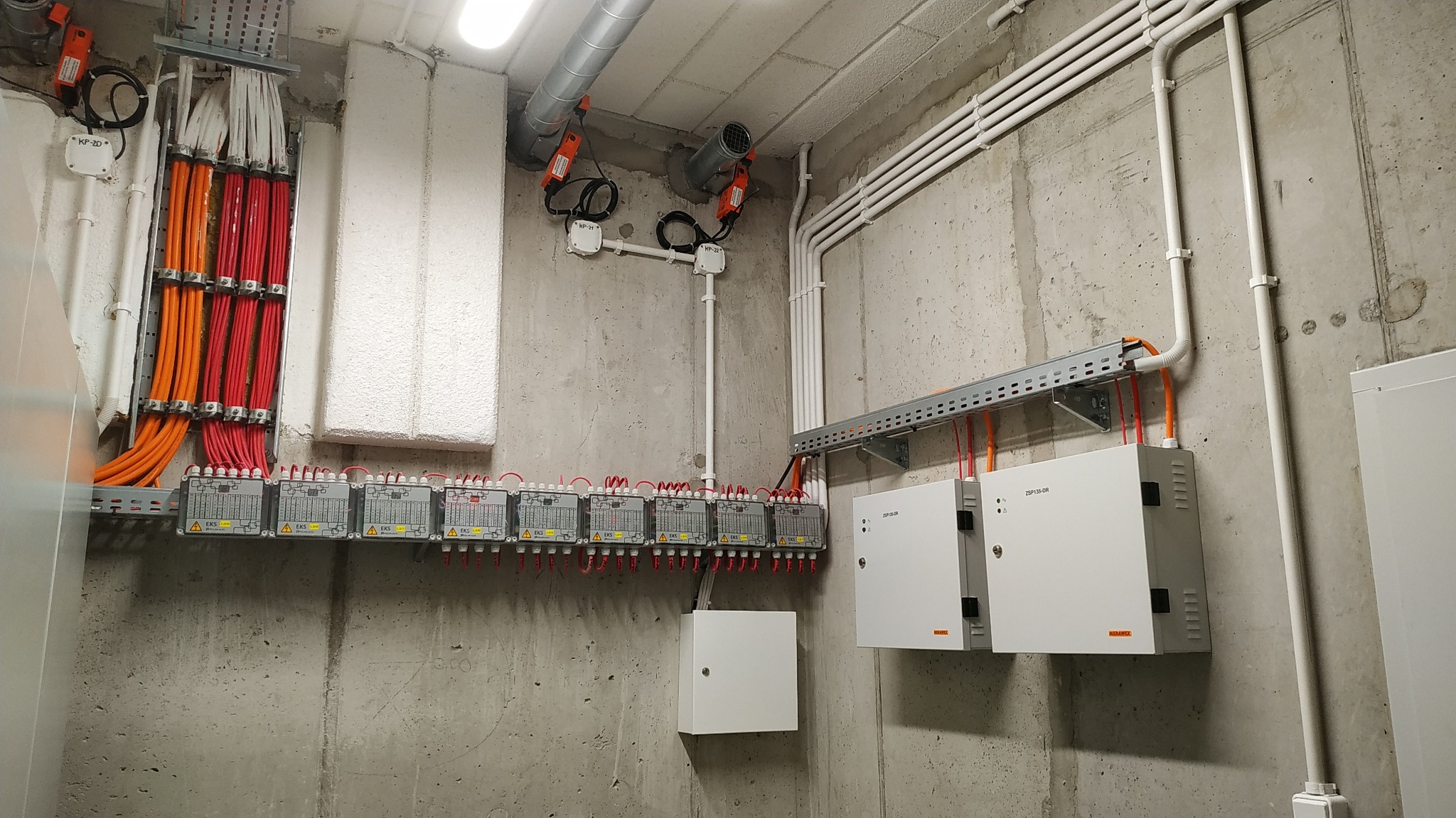 Rozbudowana instalacja elektryczna w Krakowie, z licznymi przewodami w kolorach czerwonym i pomarańczowym, prowadzącymi do szeregu modułów EKS zamontowanych na szynach, oraz białe kable w peszlach,...