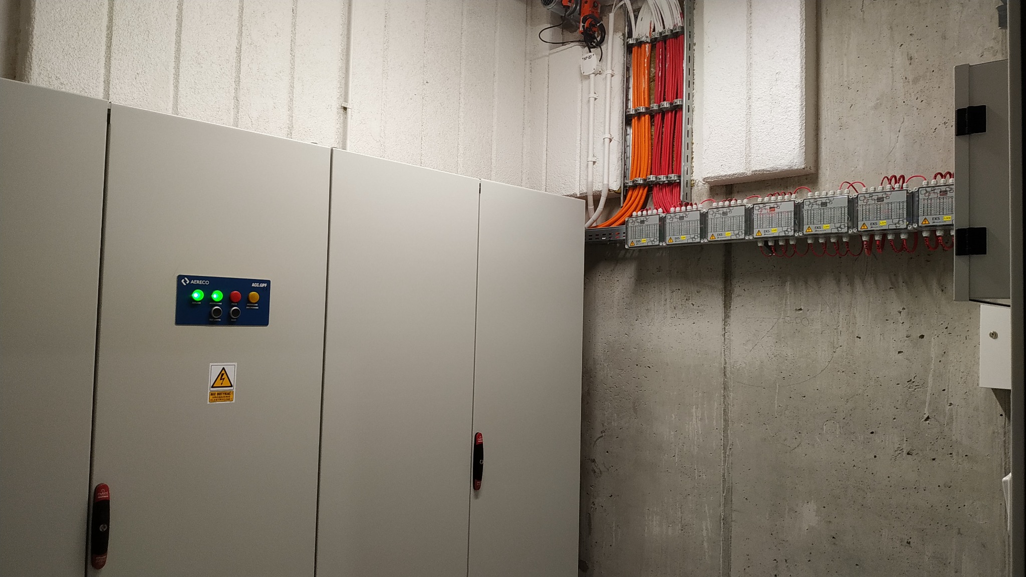 Szafa sterownicza z widocznymi przewodami w kolorze czerwonym i pomarańczowym, ułożonymi w korytkach na szarej, betonowej ścianie. Widoczne moduły EKS z czerwonymi przewodami. Na szafie panel...
