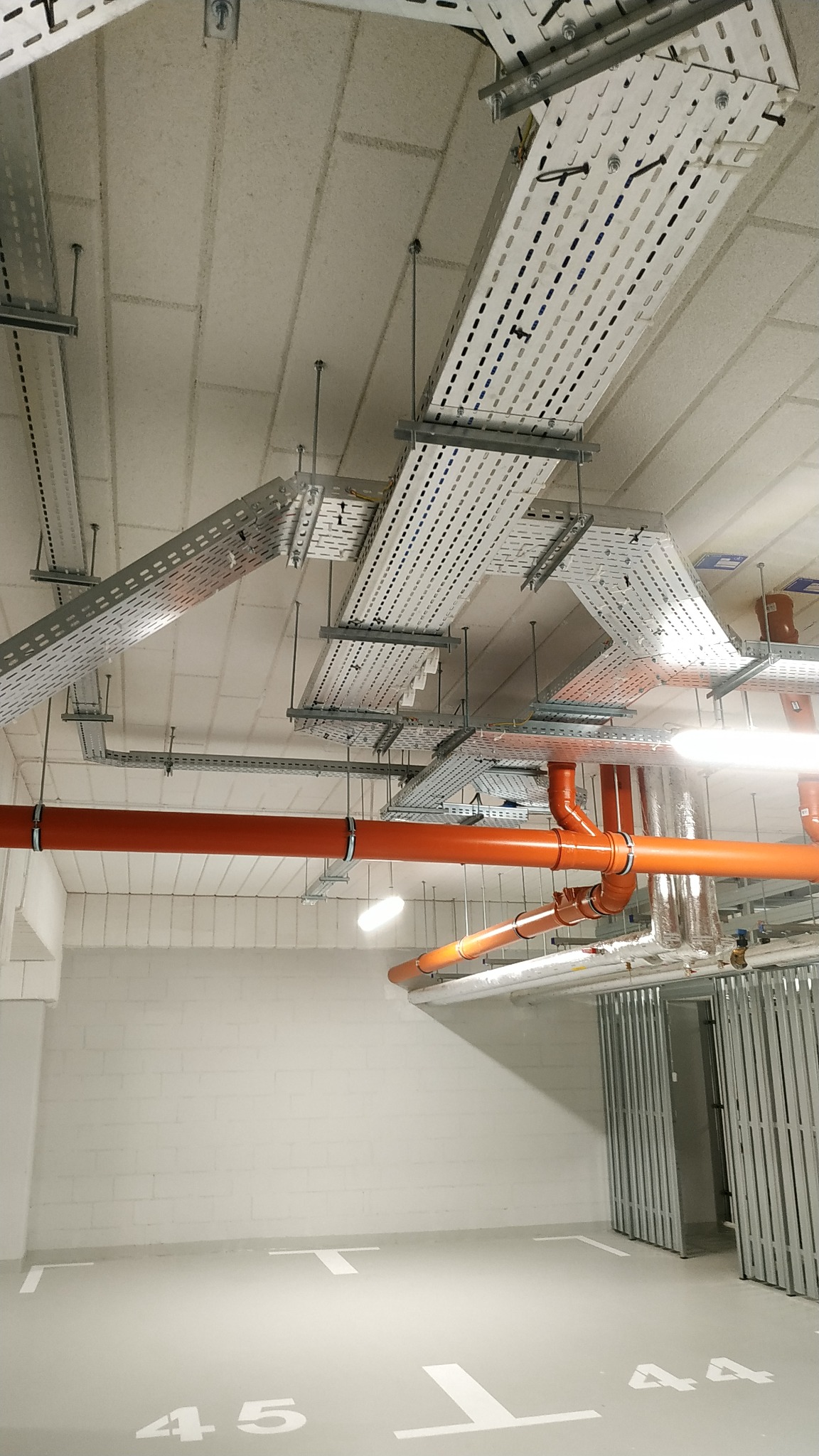 Białe koryta kablowe pod sufitem parkingu podziemnego, pomarańczowe rury wentylacyjne, białe oznaczenia miejsc parkingowych na szarej posadzce.