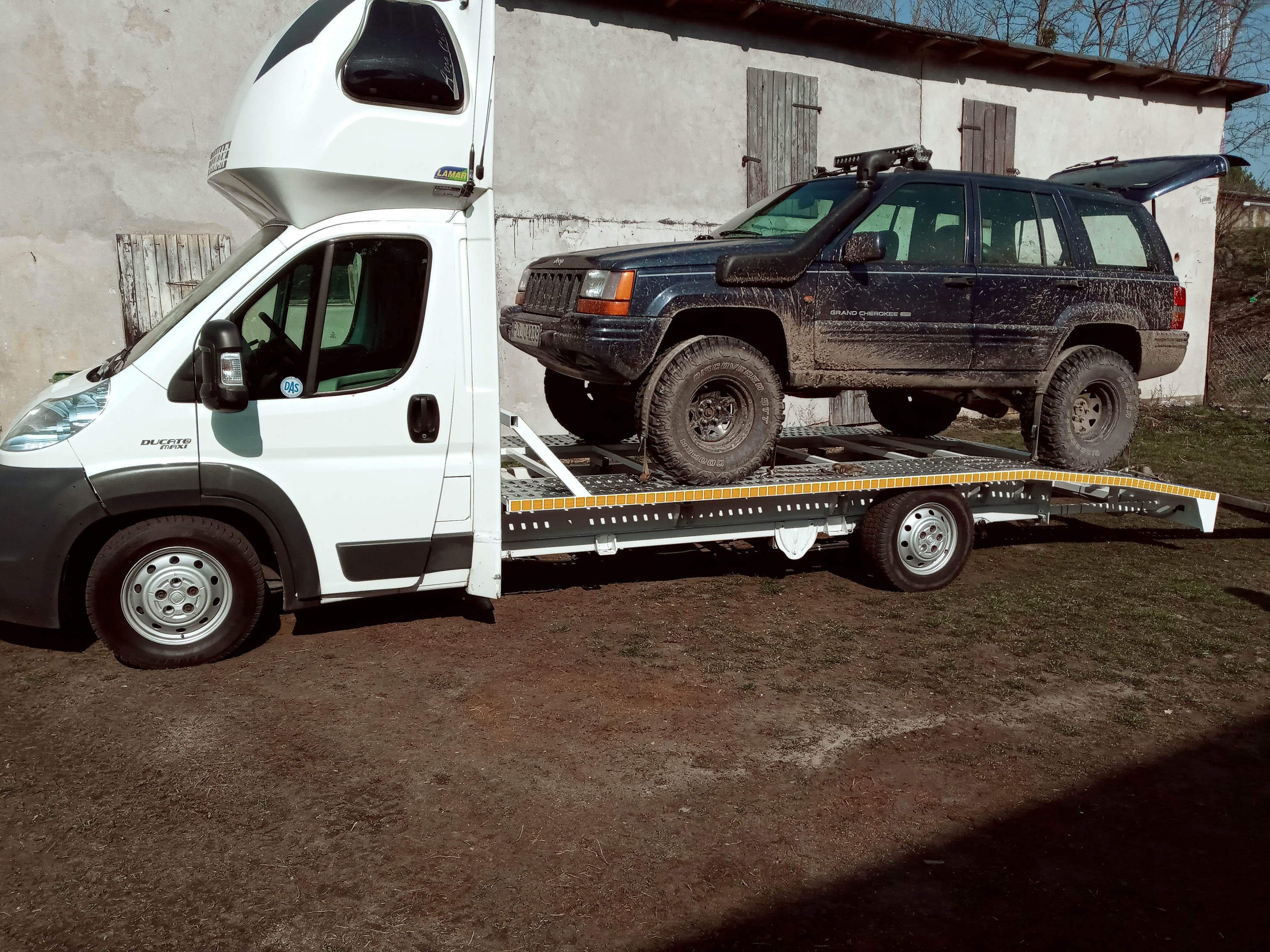 Biały bus z platformą transportową przewozi brudny, czarny samochód terenowy Jeep Grand Cherokee z otwartą klapą bagażnika na tle szarego budynku gospodarczego z drewnianymi okiennicami.
