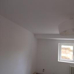 Pol Bud - Świeżo pomalowane na biało wnętrze pokoju z widocznym oknem, grzejnikiem i lampą sufitową, zabezpieczone folią malarską, przygotowane do dalszych prac wykończeniowych.