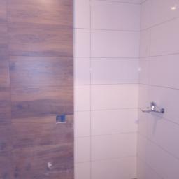 Pol Bud - Układanie płytek w łazience: ściana wyłożona płytkami drewnopodobnymi sąsiaduje z białą ścianą z zamontowaną baterią. Na podłodze folia ochronna i worek z zaprawą.