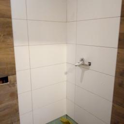 Pol Bud - Fragment łazienki w trakcie prac wykończeniowych: białe, kwadratowe płytki ceramiczne na ścianach, brązowe płytki imitujące drewno po bokach, zabezpieczona podłoga folią, widoczna bateria umywalkowa.