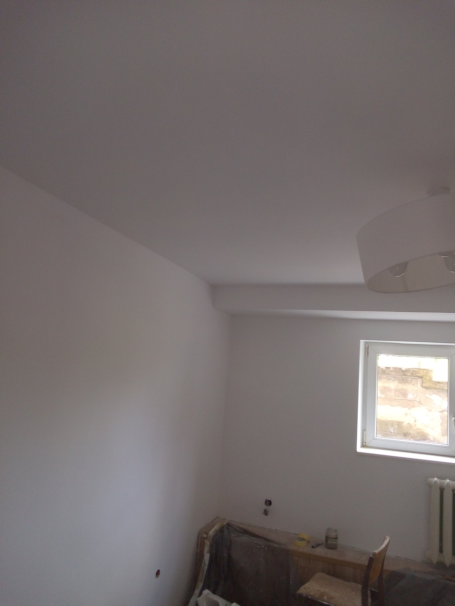 Świeżo pomalowane na biało wnętrze pokoju z widocznym oknem, grzejnikiem i lampą sufitową, zabezpieczone folią malarską, przygotowane do dalszych prac wykończeniowych.