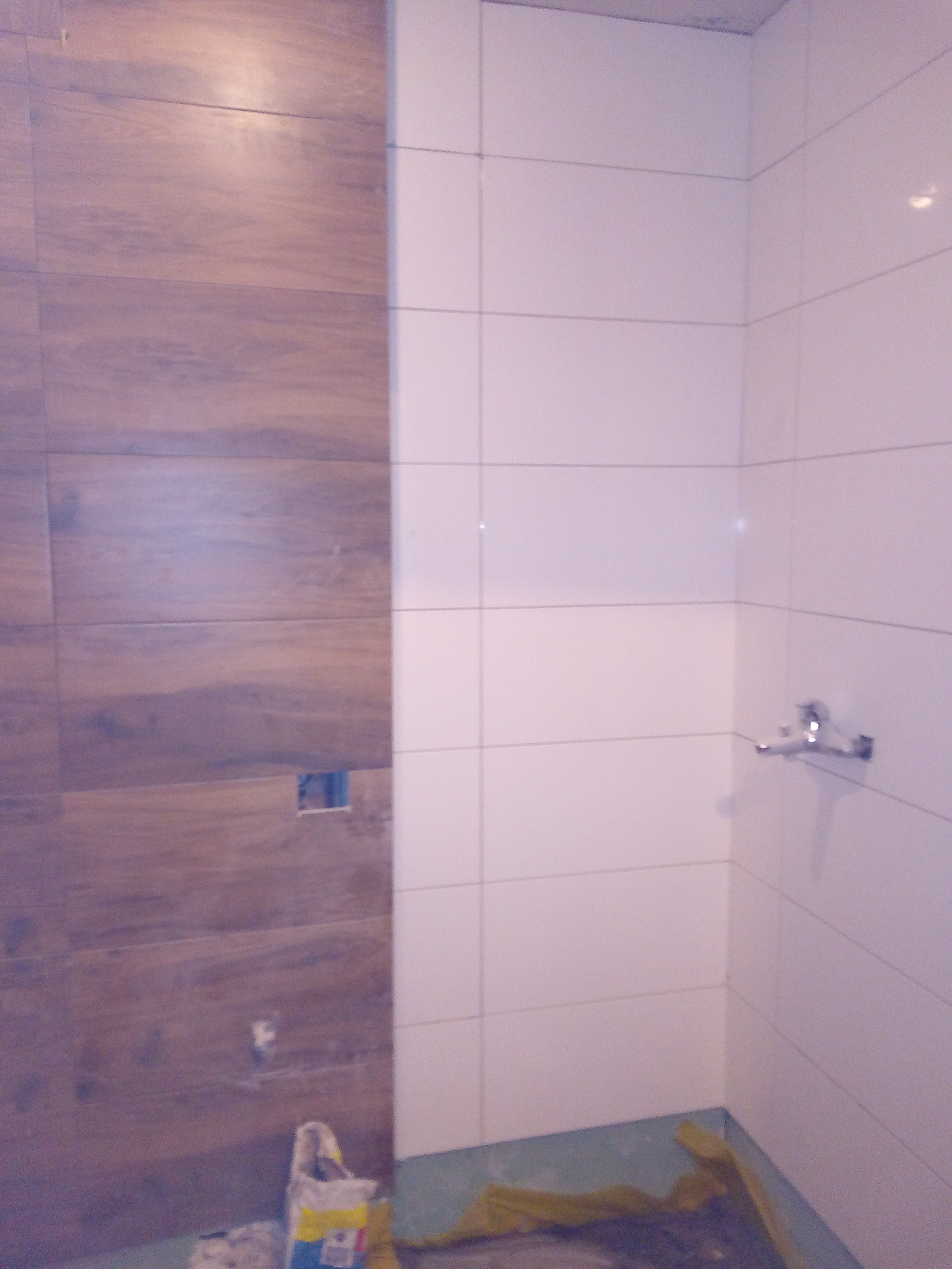 Układanie płytek w łazience: ściana wyłożona płytkami drewnopodobnymi sąsiaduje z białą ścianą z zamontowaną baterią. Na podłodze folia ochronna i worek z zaprawą.