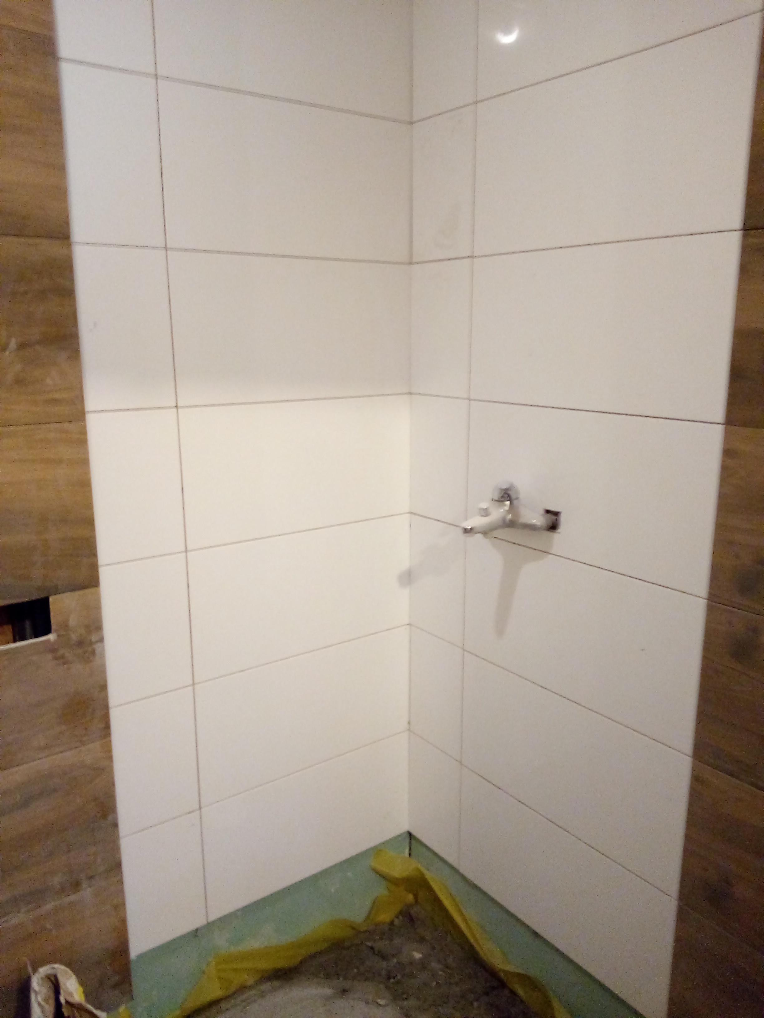 Fragment łazienki w trakcie prac wykończeniowych: białe, kwadratowe płytki ceramiczne na ścianach, brązowe płytki imitujące drewno po bokach, zabezpieczona podłoga folią, widoczna bateria umywalkowa.