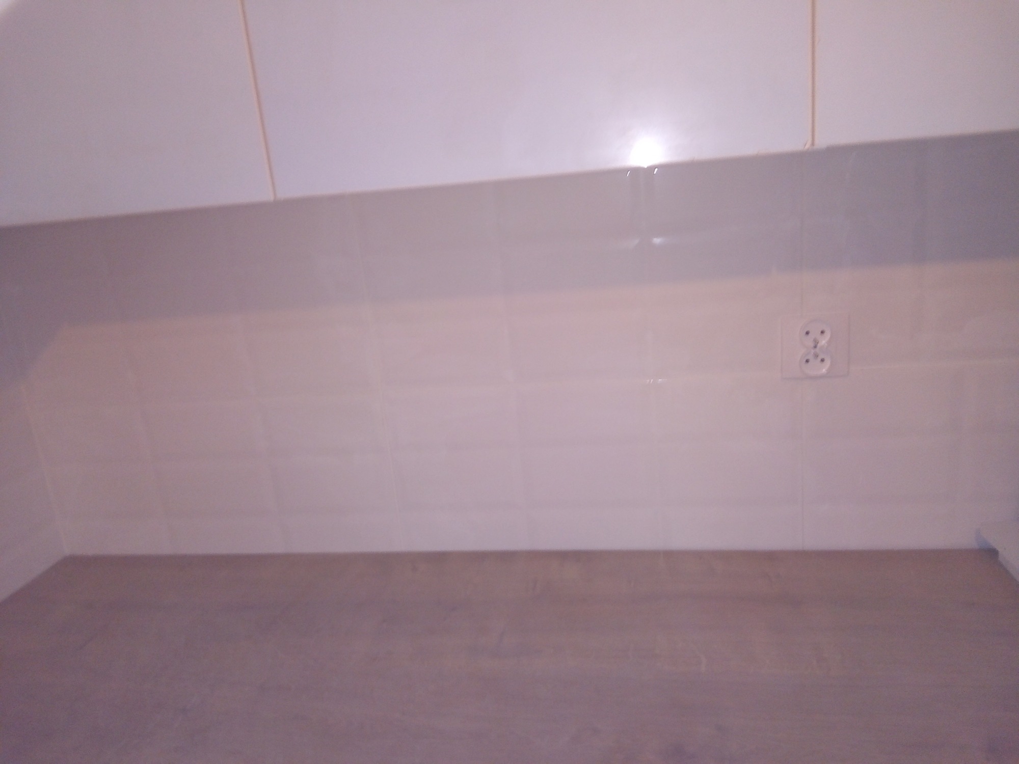 Białe płytki ścienne z widocznym gniazdkiem elektrycznym i fragmentem szafek kuchennych nad blatem.