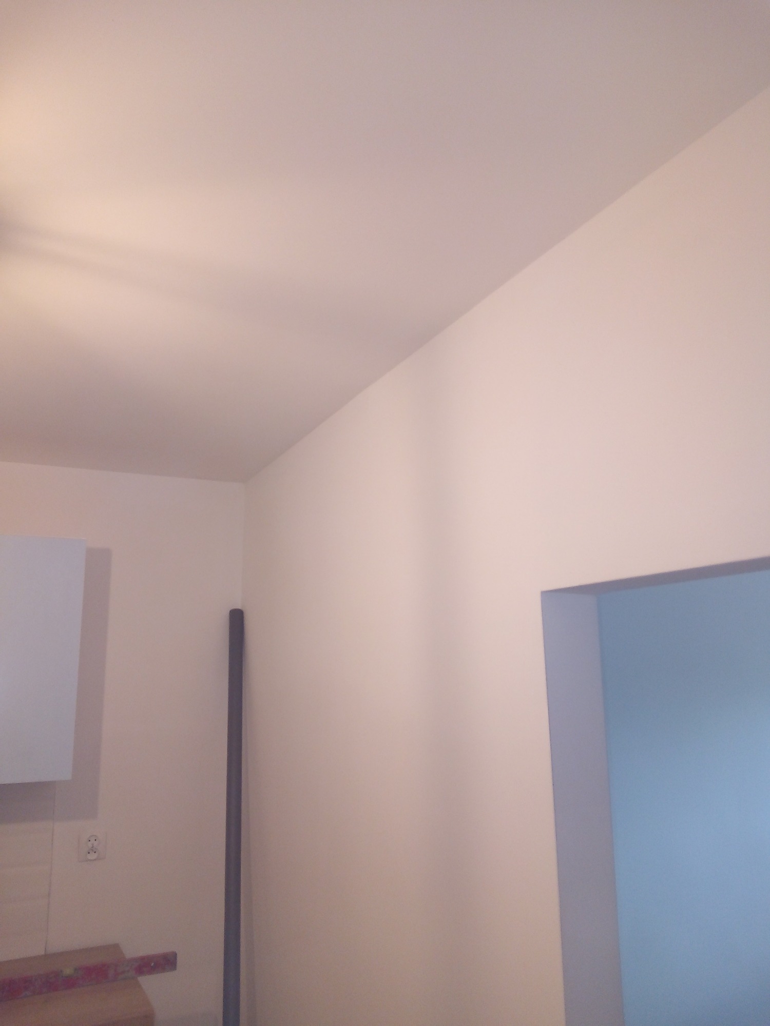 Świeżo pomalowane ściany w jasnym odcieniu beżu, widoczny fragment białej szafki, pionowa rura i fragment drzwi do innego pomieszczenia pomalowanego na niebiesko.