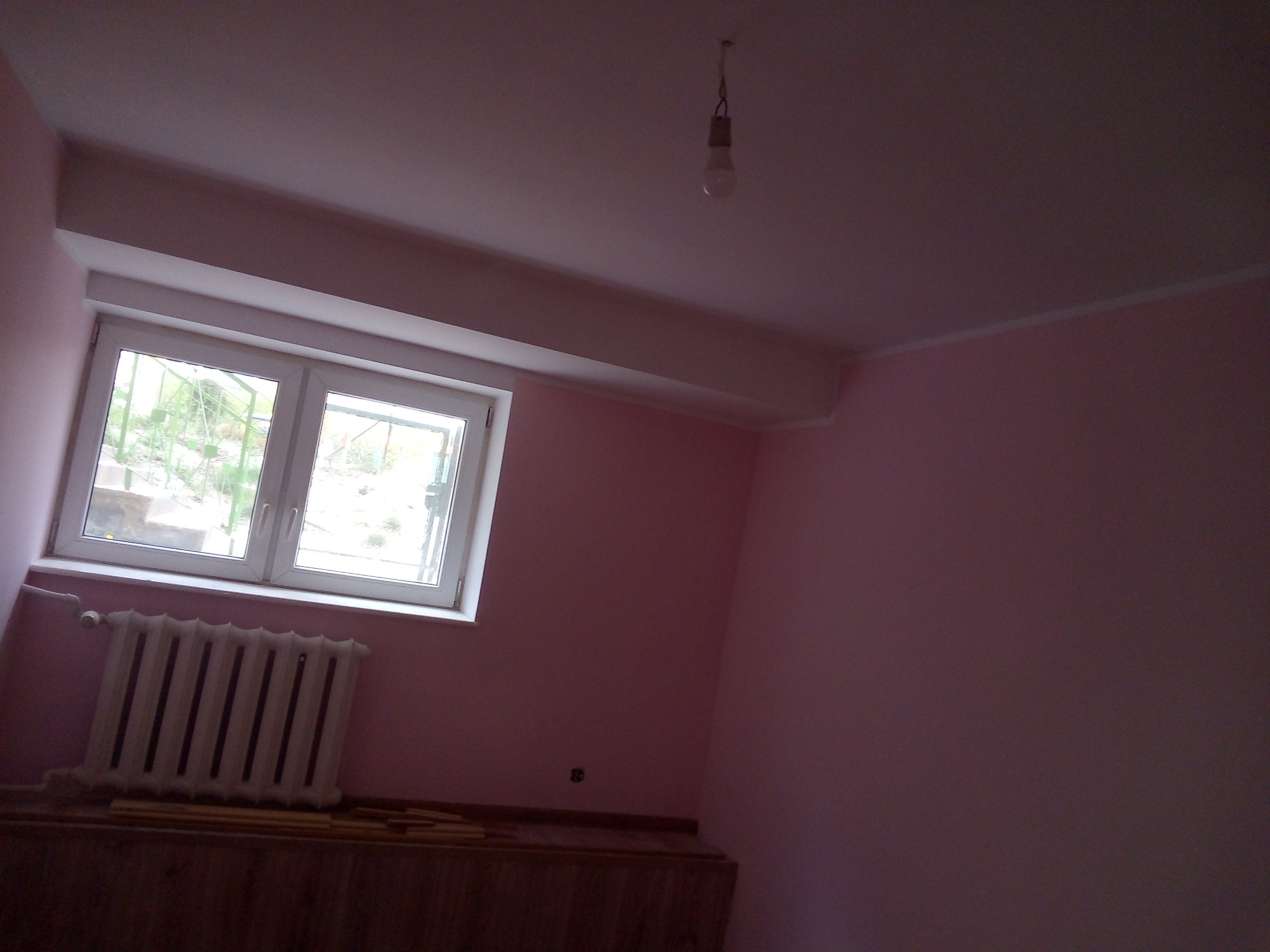Pokój po malowaniu na różowo, widoczny grzejnik, okno i surowa żarówka na kablu.