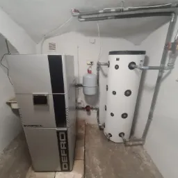 Kocioł na paliwo stałe Defro Evopell i biały zasobnik ciepłej wody w kotłowni. Widoczne rury instalacji CO i CWU.