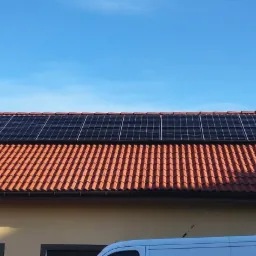 Panele słoneczne na pomarańczowym dachu domu jednorodzinnego, błękitne niebo w tle, widoczny fragment białego samochodu dostawczego.