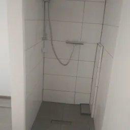 Nowoczesna kabina prysznicowa z odpływem liniowym, wykończona białymi, kwadratowymi płytkami na ścianach i szarą, kwadratową podłogą. Widoczne rury doprowadzające wodę.