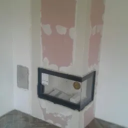 Narożny kominek z czarną ramą w trakcie obudowy płytami kartonowo-gipsowymi, widoczne ślady szpachlowania i wykończona podłoga z płytek ułożonych w jodełkę.