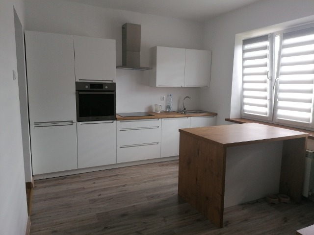 Jasna kuchnia z białymi szafkami, drewnianym blatem i wyspą kuchenną, widok z niskiego kąta, rolety w oknie.