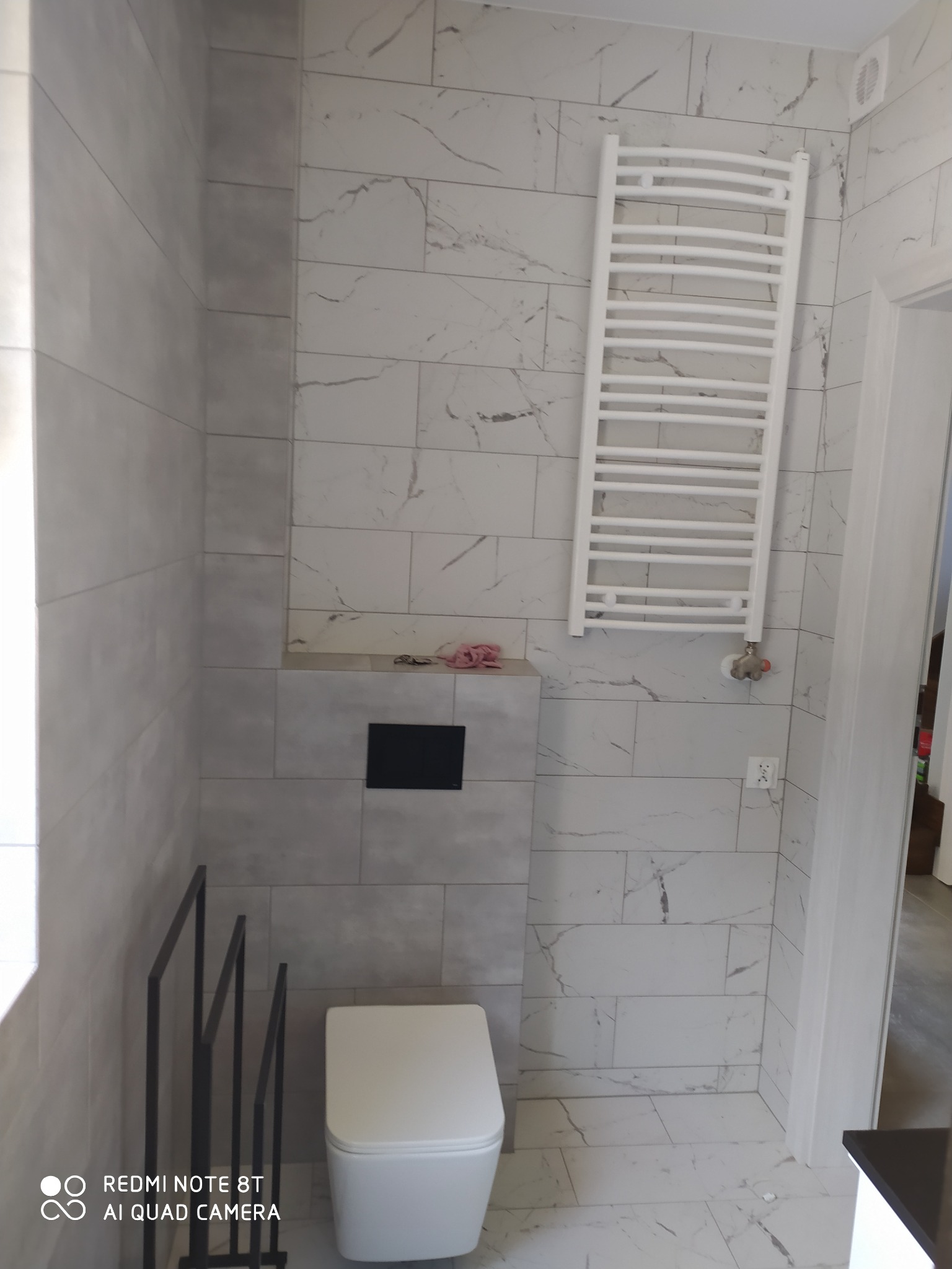 Wykończona łazienka z białymi płytkami imitującymi marmur, wiszącą toaletą, grzejnikiem drabinkowym i czarnym wieszakiem na ręczniki.