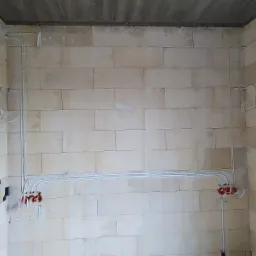 Surowa ściana z betonu komórkowego z widocznymi, nowo położonymi przewodami elektrycznymi w peszlach oraz puszkami instalacyjnymi w kolorze pomarańczowym.