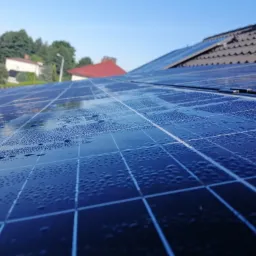 Bliższe ujęcie paneli słonecznych na dachu domu, pokrytych drobnymi kroplami wody, z widocznym fragmentem dachu z dachówki i błękitnym niebem w tle.