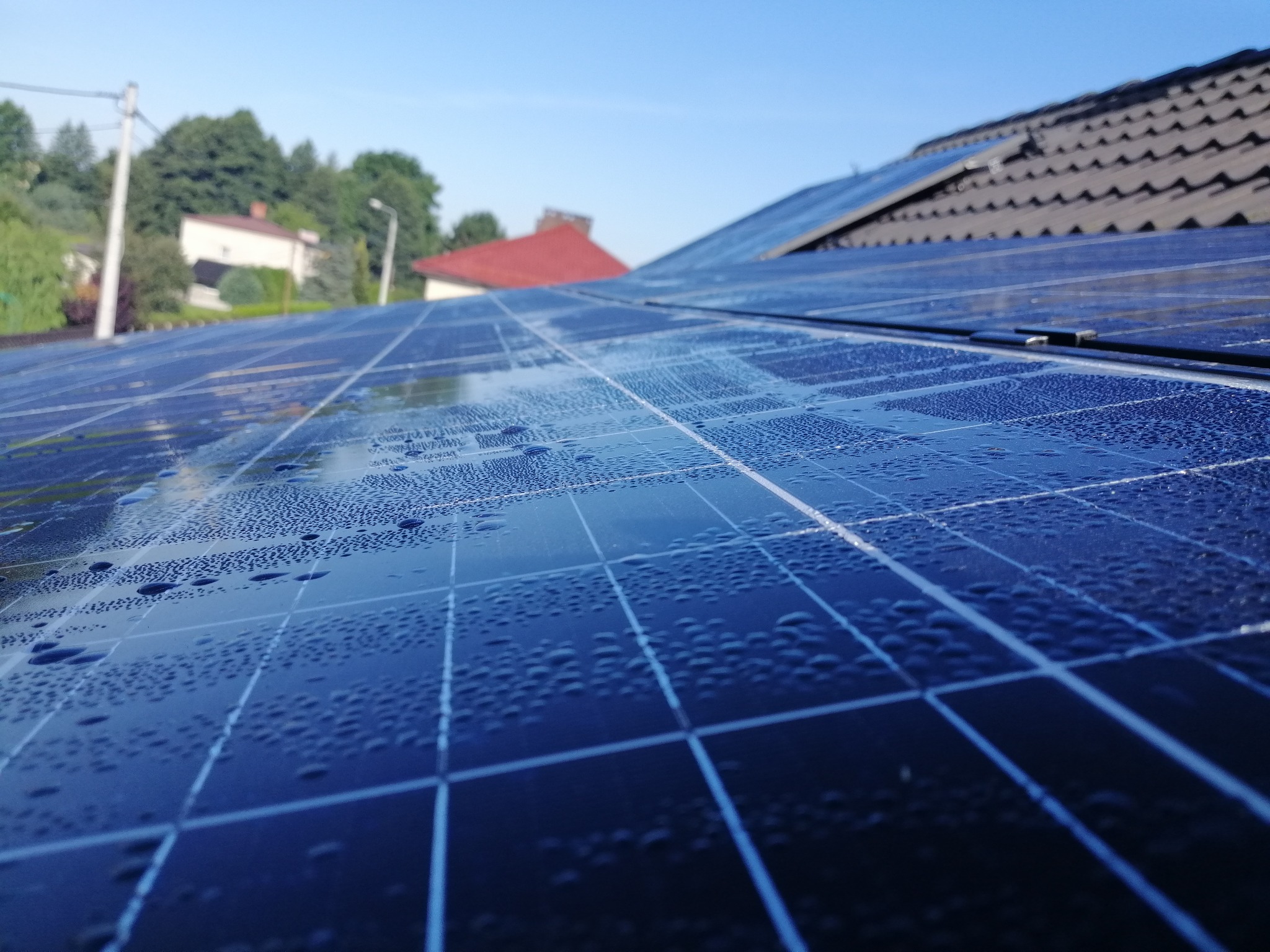 Bliższe ujęcie paneli słonecznych na dachu domu, pokrytych drobnymi kroplami wody, z widocznym fragmentem dachu z dachówki i błękitnym niebem w tle.