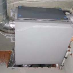 Szara centrala rekuperacyjna montowana pod sufitem, widoczne rury wentylacyjne, instalacja w trakcie prac wykończeniowych.