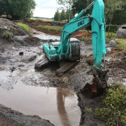 Turkusowa koparka Kobelco zanurzona w błotnistym wykopie, odbicie maszyny w wodzie, w tle zalesiony teren i druga koparka na wzgórzu.