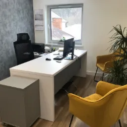 Nowoczesne biuro: białe biurko z komputerem, żółte krzesła welurowe, szara ściana z fakturą, widok na zimowy krajobraz za oknem, kalendarz na ścianie.