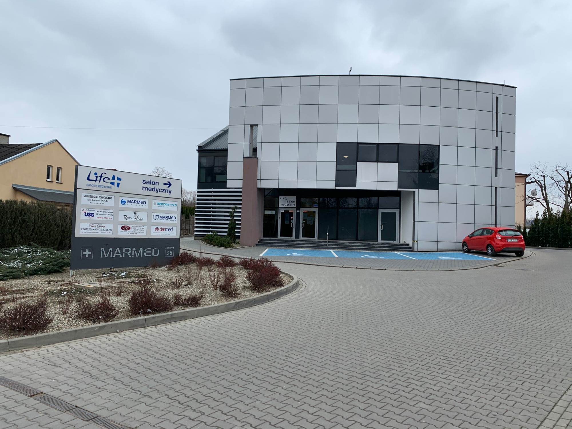 Nowoczesny budynek medyczny z szarą elewacją panelową i dużymi oknami, przed budynkiem oznakowany parking dla osób niepełnosprawnych i tablica informacyjna z nazwami firm i usług medycznych.