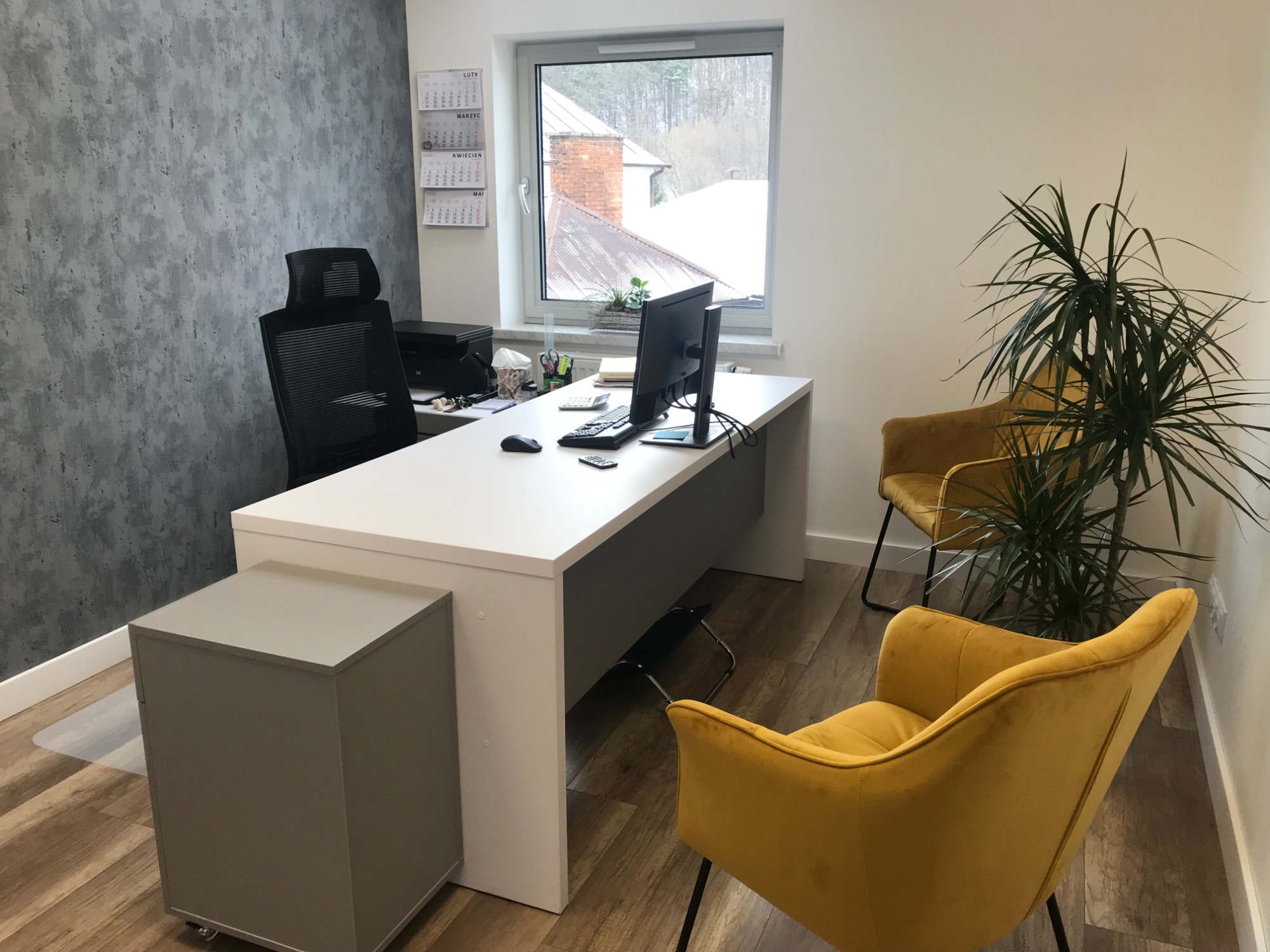 Nowoczesne biuro: białe biurko z komputerem, żółte krzesła welurowe, szara ściana z fakturą, widok na zimowy krajobraz za oknem, kalendarz na ścianie.